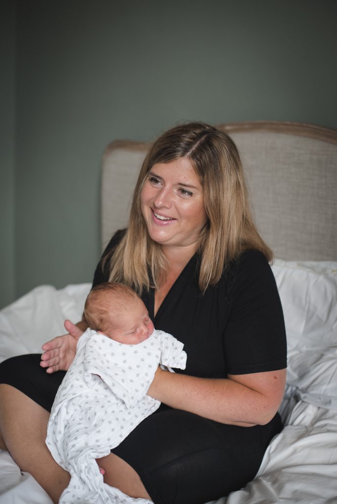 An image of Tricia Murray. She is a white woman with blond hair wearing a black dress. She is sat on a bed and holding a very small baby