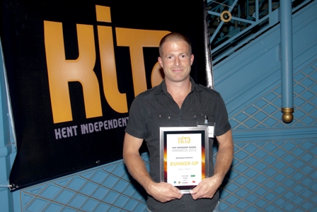 An image of Ben Smith. He is a man with very short hair. He is wearing a grey top and is holding a certificate in a frame and he is stood in front of a banner.