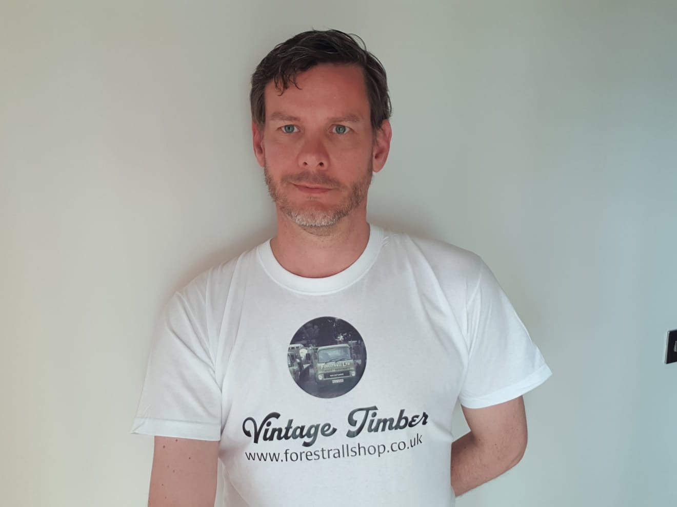 An image of Chris Porcas. He is a man with brown hair and some facial hair. He is wearing a white t-shirt with a logo for Vintage Timber and a web address.