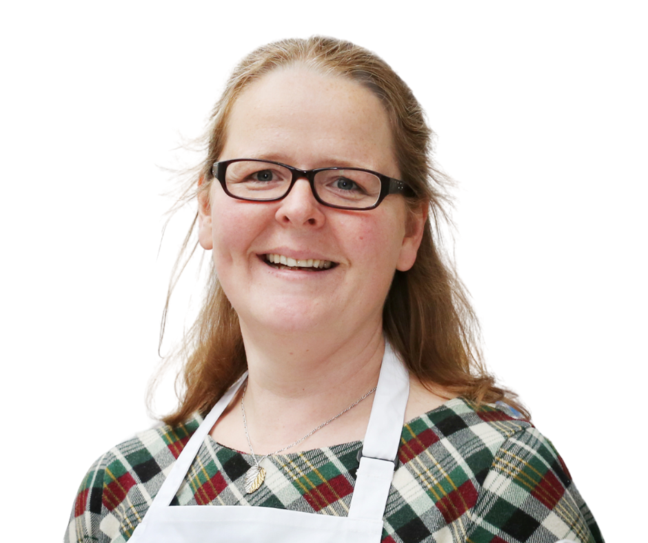 An image of Hadi Brooks. She is a white woman with ginger hair and glasses. She is wearing a tartan dress and a white apron. The image is a headshot and she is smiling at the camera.