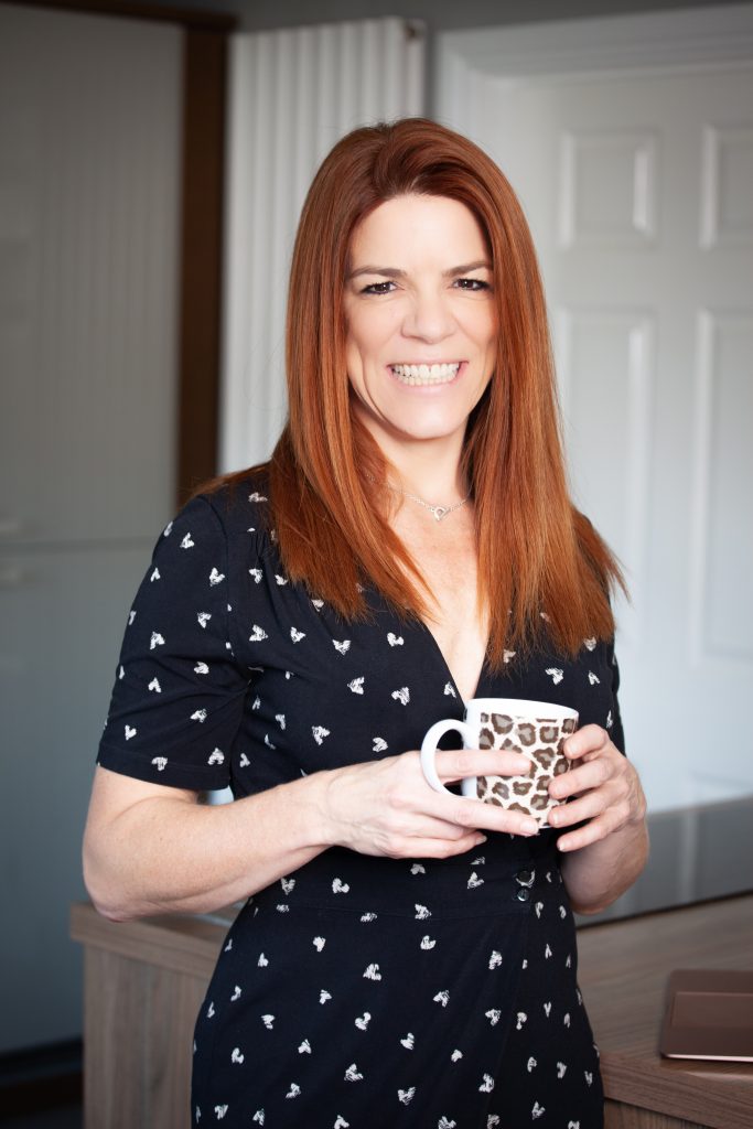 An image of Jo Francis. She is a white woman with ginger hair wearing a navy blue dress and holding a leopard print mug.