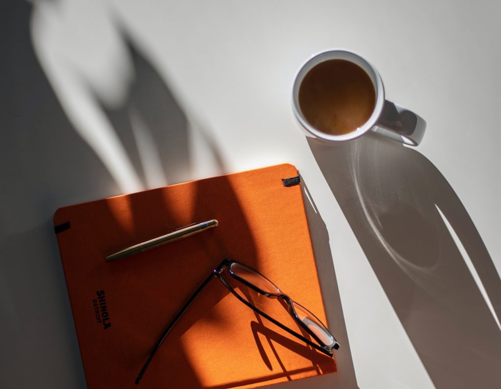 An image of a table with shadows cast over it. On the table there is a full mug of tea and an orange notebook with a pair of glasses and a pen resting on top of it.