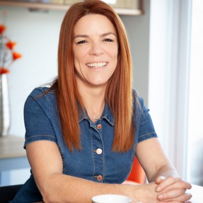An image of Jo Francis who is a white woman with ginger hair wearing a blue denim short sleeved blouse sat at a table with a leopard print mug in front of her clasped hands.